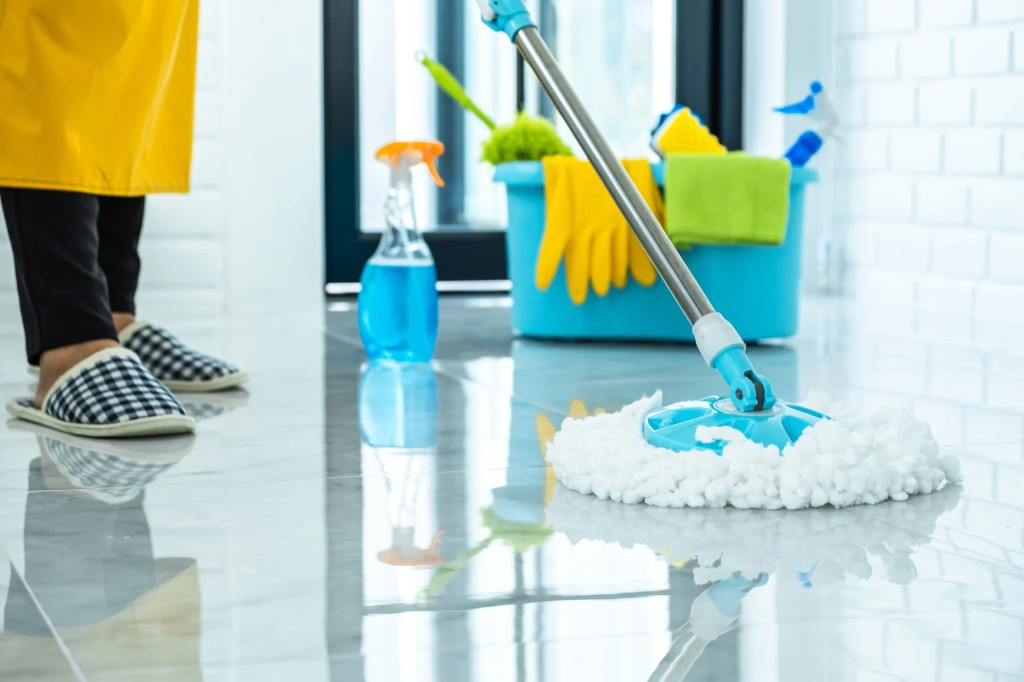Cleaner mopping a polished tile floor with a blue bucket of supplies nearby